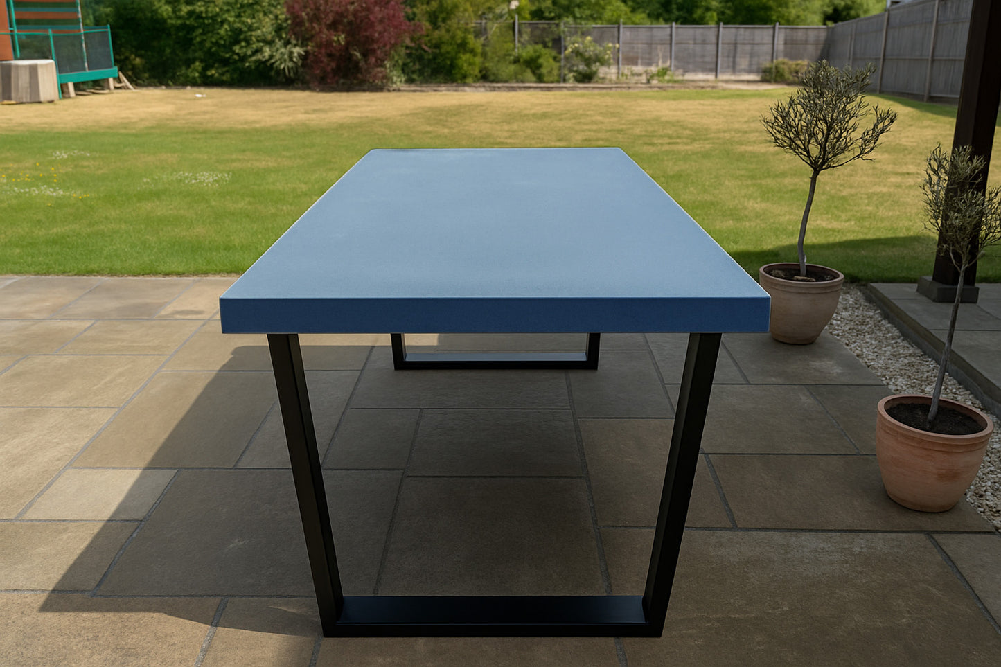 Blue table with black legs on a patio area with potted plants and grass in the background