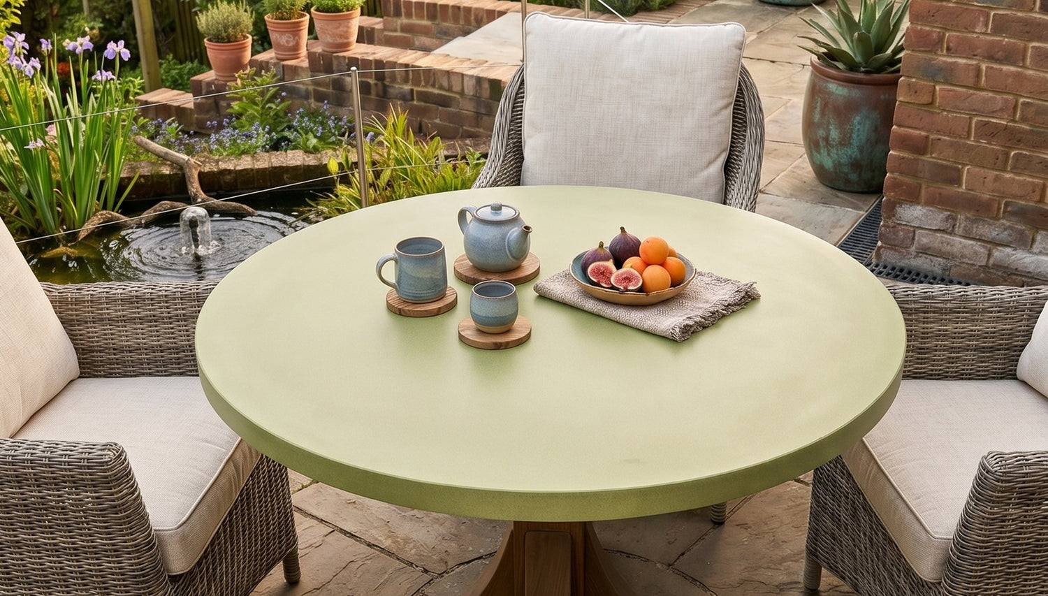 Round green concrete table with teacups and fruit plate in a garden setting with wicker chairs and potted plants.