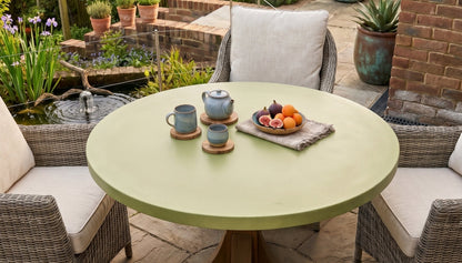 Round green concrete table with teacups and fruit plate in a garden setting with wicker chairs and potted plants.